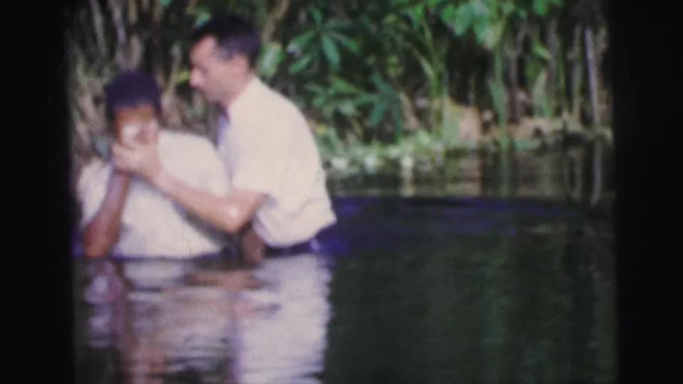 1967 Brazil 8mm frame of an apparent outdoor baptism, with one man supporting another in shallow water.