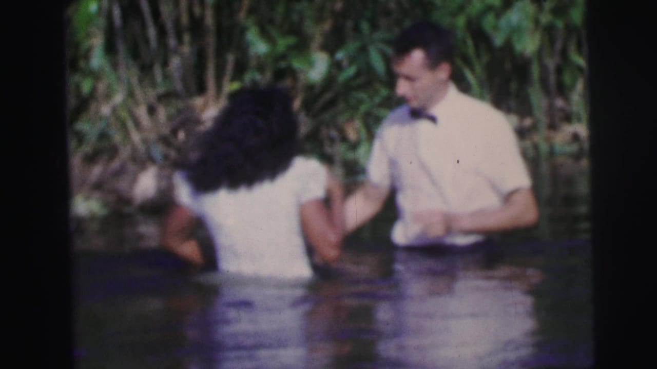 Woman and man stand waist-deep in water beside dense foliage during a baptismal moment.