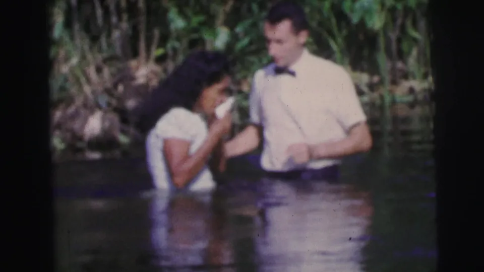 The woman appears to hold or cover her mouth as the man stands beside her in a tropical waterside baptism scene in Brazil.