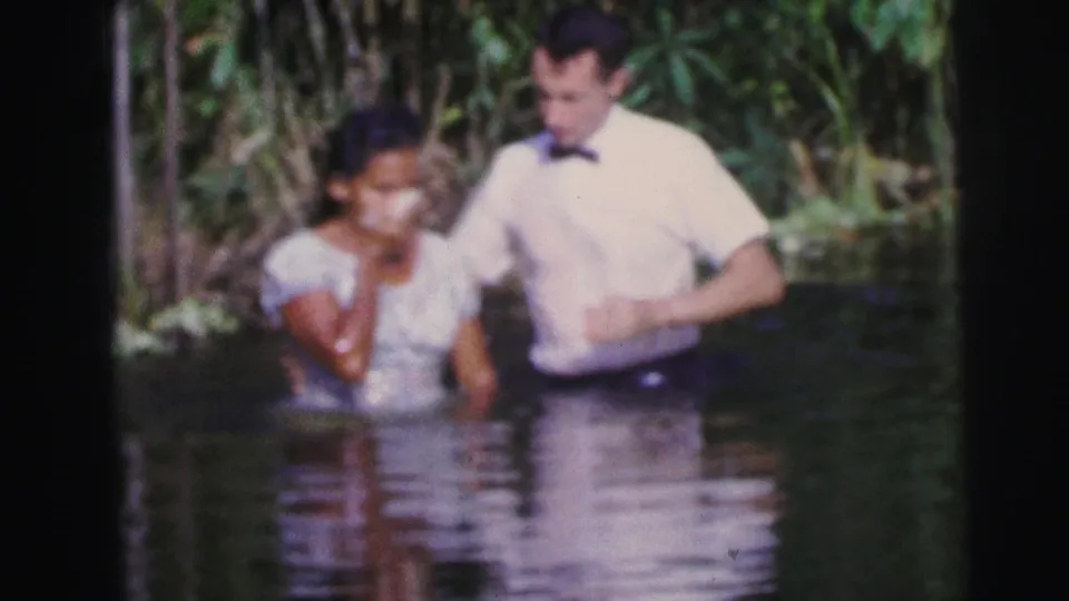 The woman stands beside the man in shallow water, still holding a hand near her mouth, in a 1967 Brazil outdoor baptism scene.