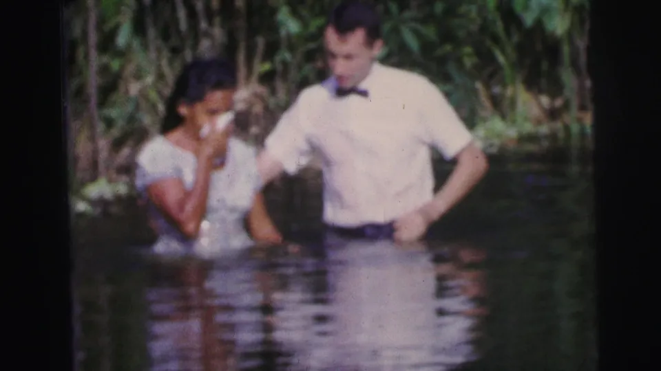 In 1967 Brazil home-movie footage, a woman holds a cloth near her face while standing in shallow water beside a man in formal dress.