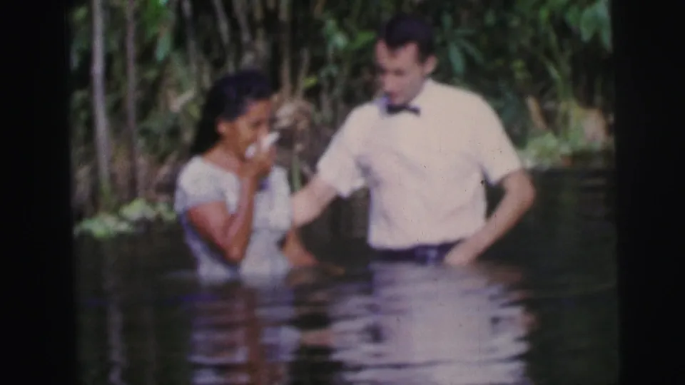 The man appears to guide or support the woman as she holds a cloth to her face in shallow outdoor water, with greenery behind them.