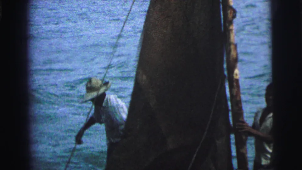 1967 Brazil 8mm frame with a hat-wearing man leaning near rigging while a dark sail fills the right side of the shot.
