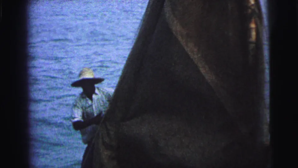 1967 Brazil home-movie frame showing a man in a straw hat near the sail, with rigging and ocean water behind him.