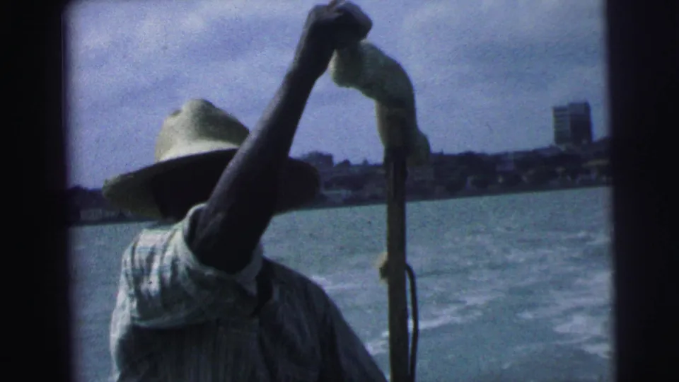 1967 Brazil home-movie frame showing an adult man in a straw hat steering or handling a small boat, with water and city buildings behind him.
