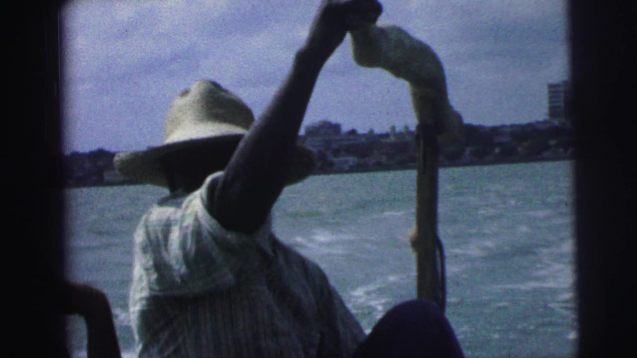 1967 Brazil 8mm frame of a straw-hatted man working a pole or oar from a small boat, with shoreline buildings beyond the water.