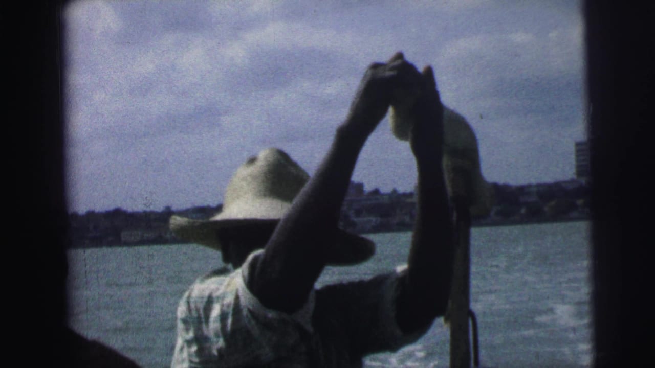 Straw-hatted man lifts his arms with a pole or oar while water and buildings sit behind him.
