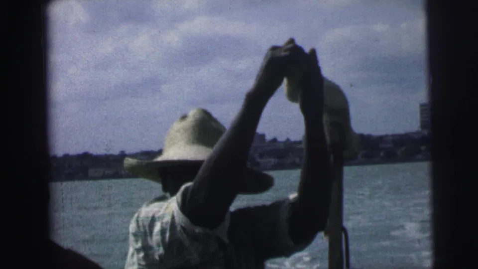 1967 Brazil home-movie frame showing a man in a straw hat handling a pole or oar on coastal water, seen from inside the boat.