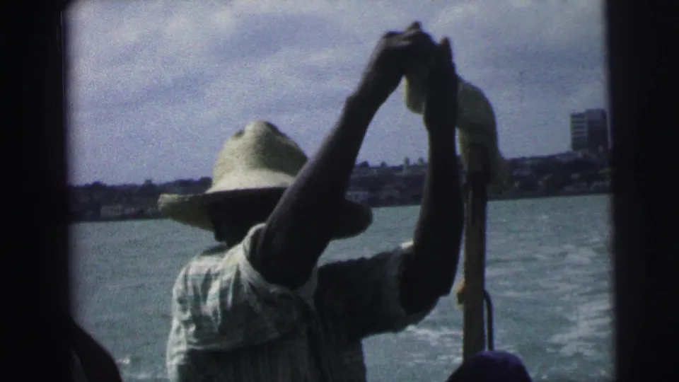 1967 Brazil 8mm frame of a boatman in a straw hat and striped shirt, partly silhouetted against water and a distant waterfront.