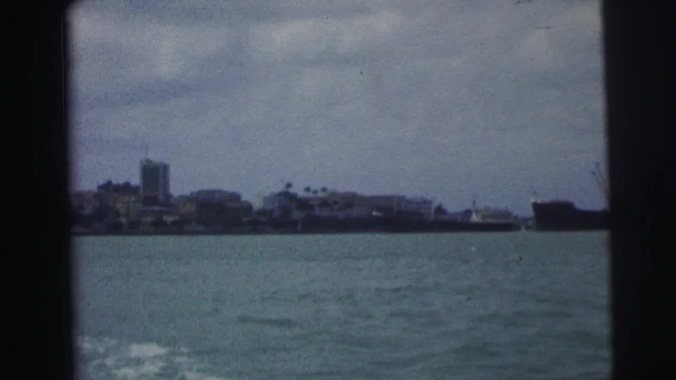 1967 Brazil home-movie frame looking across coastal water toward distant low buildings and a taller structure on the shoreline.
