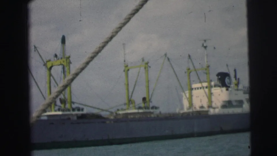 1967 Brazil 8mm frame of a large cargo vessel with yellow deck cranes, seen from a nearby moving boat.