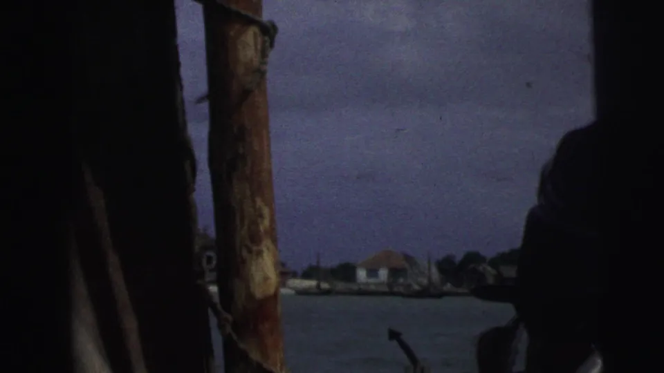 1967 Brazil 8mm frame looking from a boat toward harbor water, a distant low building, and dark foreground silhouettes.