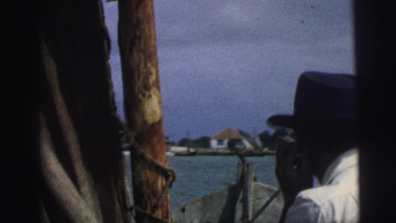 Man in a dark hat smoking or holding a cigarette on a boat, framed by wooden posts near water.