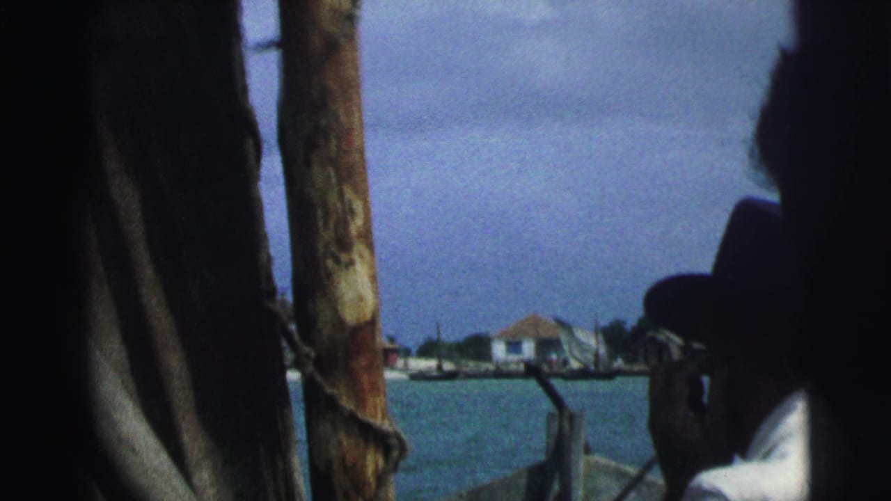 Partially obscured man in a hat on a boat with water and distant low buildings behind him.