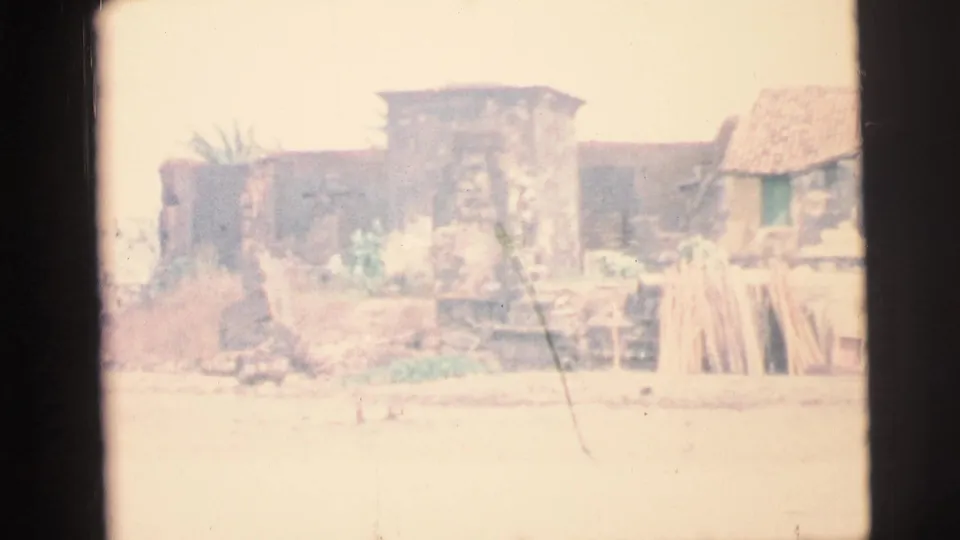 1967 Brazil 8mm frame showing a faded view of a waterside settlement with simple buildings, rooflines, and stacked wood.