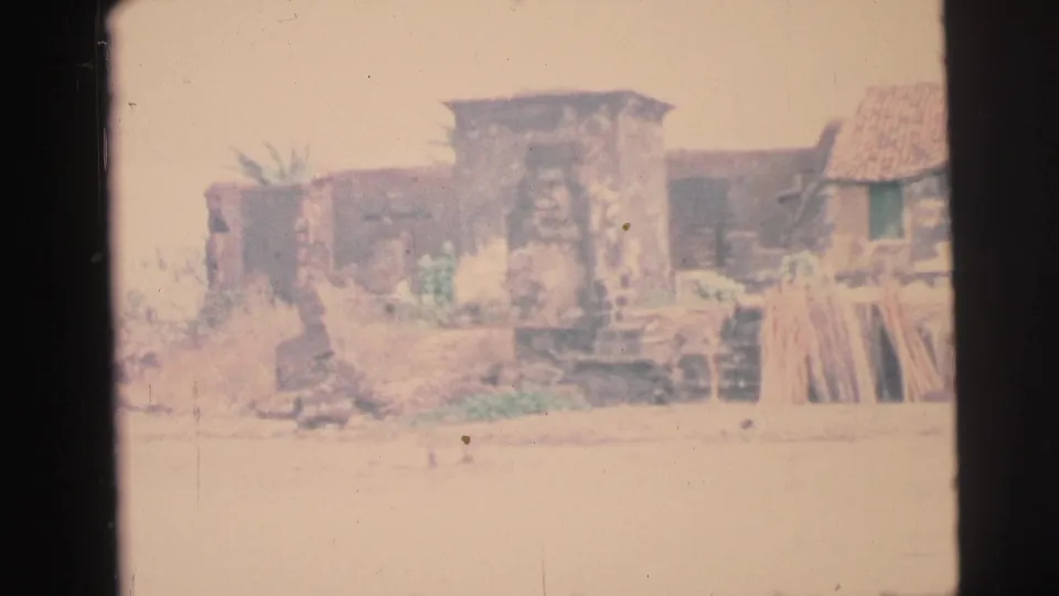 1967 Brazil home-movie frame showing low settlement buildings, a thatched or tiled roof, stacked materials, and pale open ground.