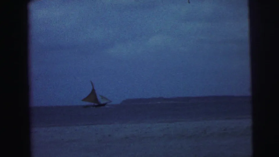 1967 Brazil 8mm frame showing a small sailboat moving across calm blue water near a low distant shore.