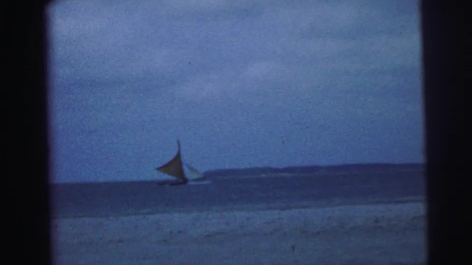 1967 Brazil home-movie frame of a small sailboat on calm coastal water beneath a muted blue sky.
