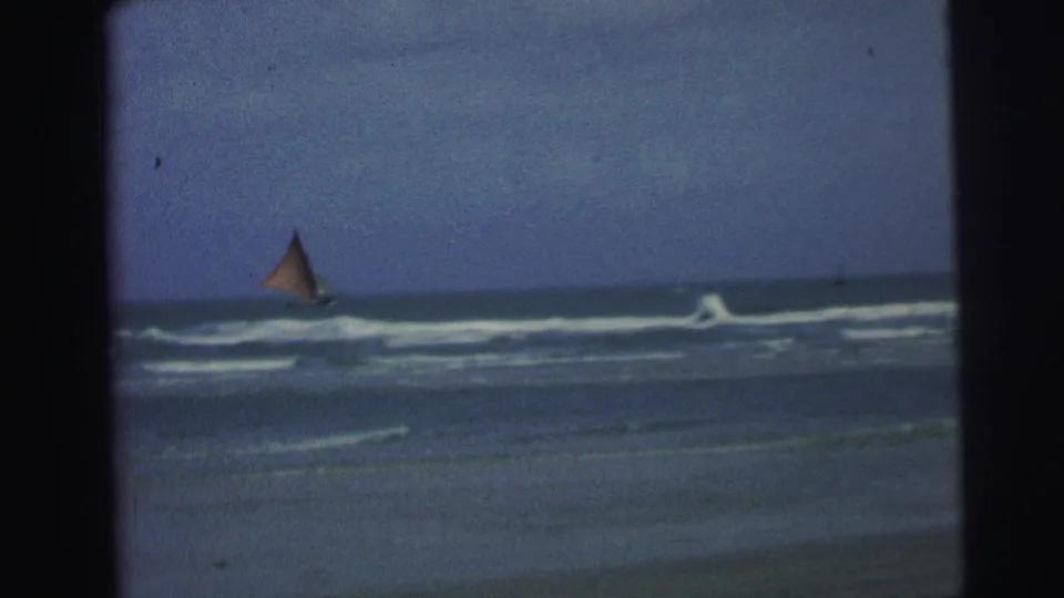 1967 Brazil 8mm frame showing a distant sailboat beyond low white surf, blue water, and a cloudy coastal sky.