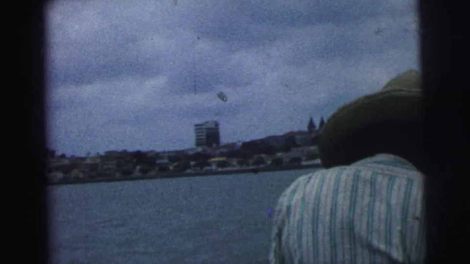 1967 Brazil 8mm frame showing a boat passenger from behind, open water, cloudy sky, and a distant urban shoreline.