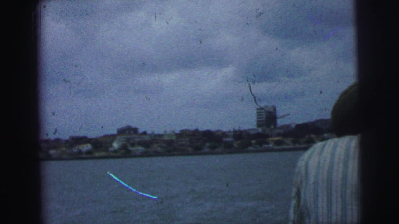 1967 Brazil 8mm frame with open water, a distant shoreline, a taller building, cloudy sky, and a partial boat passenger at right.