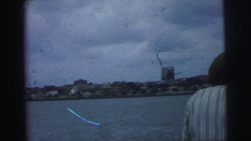 1967 Brazil 8mm frame with open water, a distant shoreline, a taller building, cloudy sky, and a partial boat passenger at right.