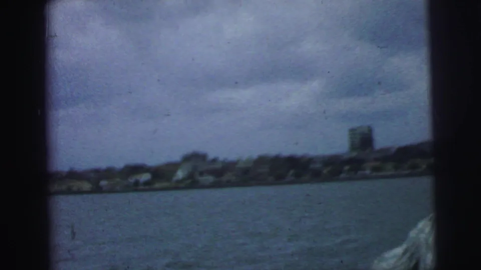 1967 Brazil 8mm frame showing a low urban shoreline across the water and a partial passenger at the edge of the boat view.