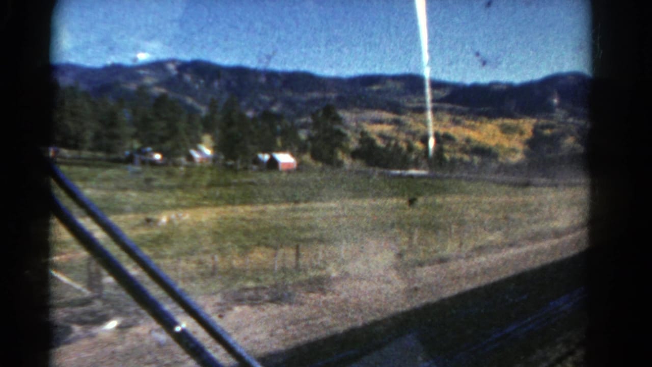 1967 Colorado rural landscape filmed from inside a moving vehicle, with fields, trees, distant hills, and windshield wipers visible.