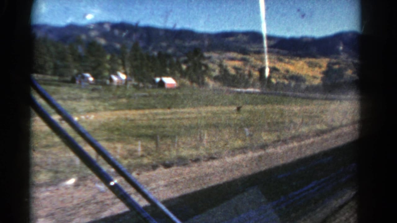 A moving-vehicle view across rural Colorado fields toward evergreen trees, low hills, and a small red farm structure in 1967 home-movie footage.