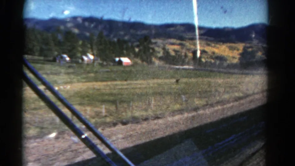 A moving-vehicle view across rural Colorado fields toward evergreen trees, low hills, and a small red farm structure in 1967 home-movie footage.