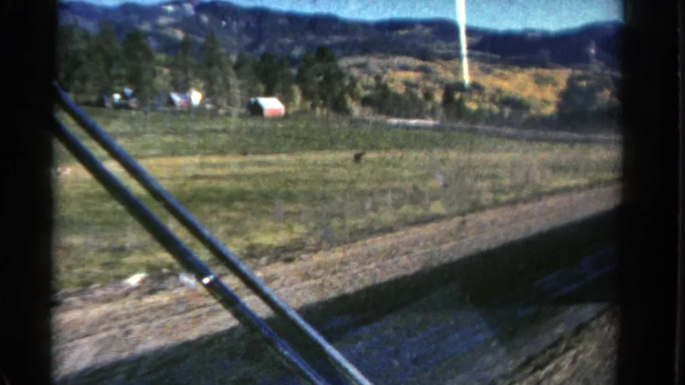 Rural Colorado landscape from a moving car, with fence posts, open field, evergreen trees, hills, and a small red building visible.