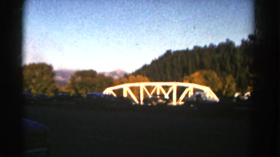 1967 Colorado 8mm frame showing a white truss bridge in the distance, bordered by trees with mountains under a blue sky.