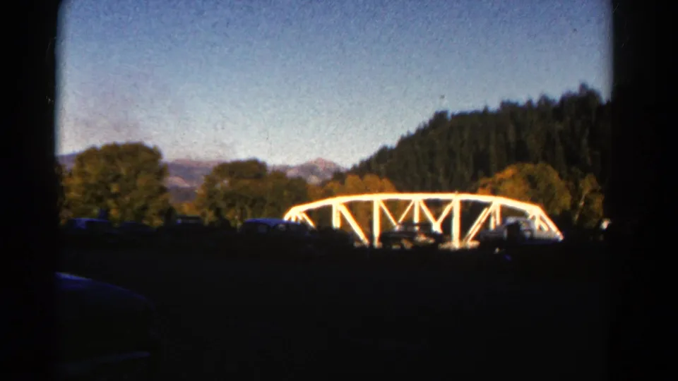 Soft 8mm Colorado travel frame showing the white bridge lower in view, with trees, mountains, blue sky, and dark vehicle-edge foreground.