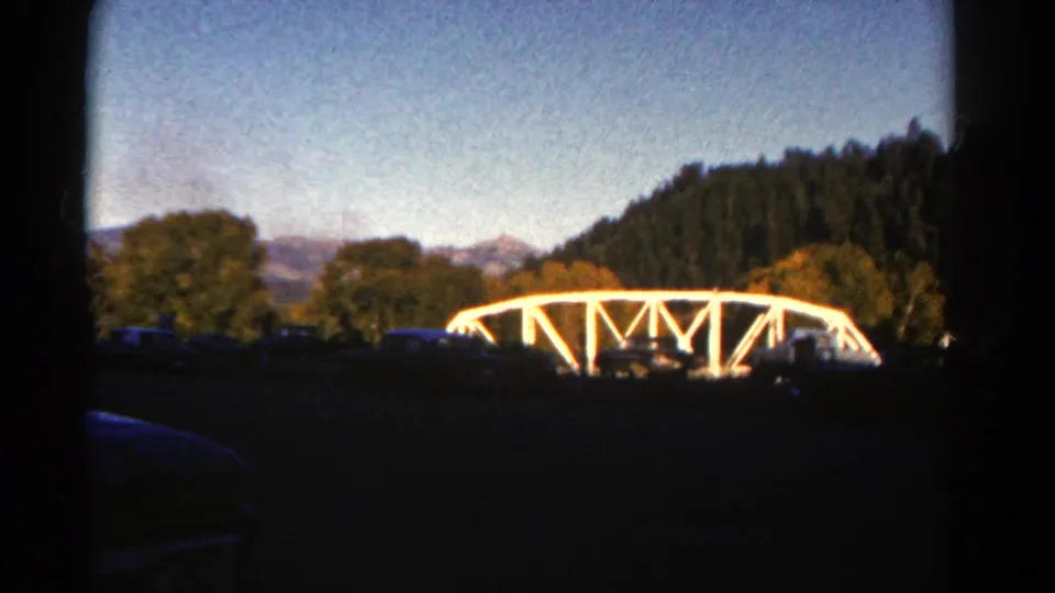 Final 1967 Colorado 8mm frame showing the white truss bridge shifted right, surrounded by trees beneath a clear mountain sky.
