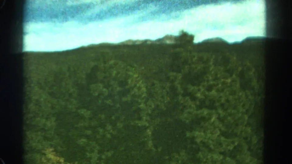 1967 Colorado home-movie frame with green treetops, a shadowed ridge, and soft sky rendered in grainy 8mm color.