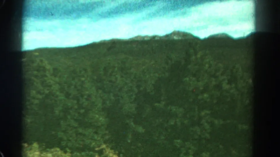 1967 Colorado 8mm frame showing a wide green forest slope with an uneven mountain horizon and bright clouded sky.