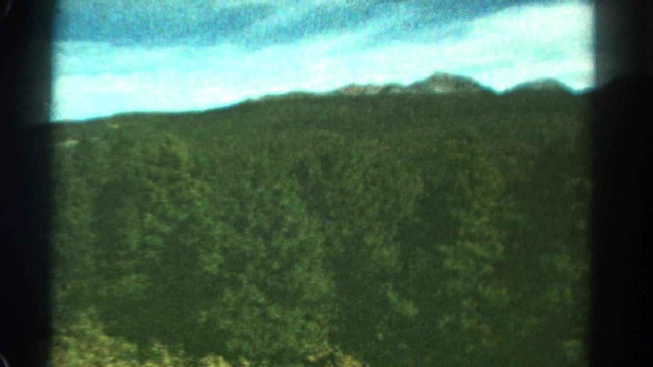 1967 Colorado 8mm frame showing green wooded hills, low mountain ridges, and cloudy blue sky with visible film gate edges.