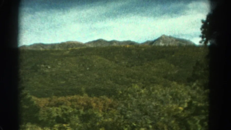 1967 Colorado 8mm landscape frame with foreground shrubs, rolling green terrain, and distant mountain ridges under clouded blue sky.