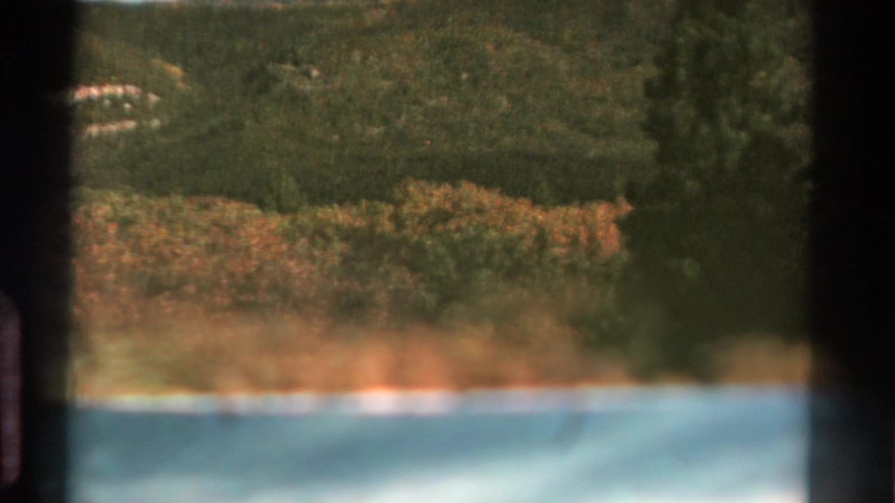 1967 Colorado home-movie frame showing water, tree-lined shore, and dark wooded hills with visible film grain and edge shadowing.