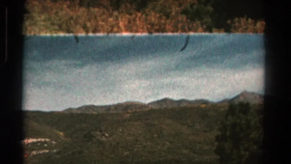 1967 8mm Colorado landscape with a band of water, wooded shoreline, rolling hills, and far mountain peaks under a pale sky.