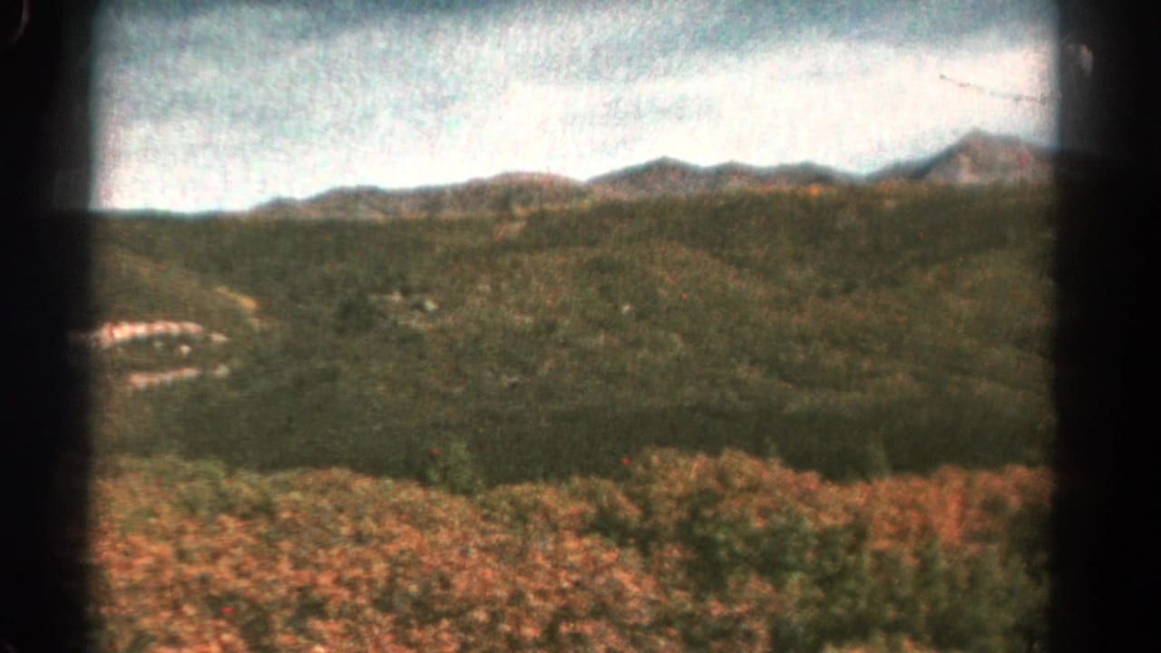 Wide 1967 Colorado landscape with rolling green hills, distant peaks, cloudy sky, and a pale winding road or clearing on the left.
