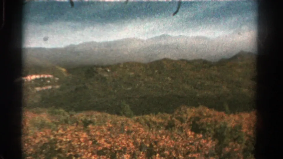 Soft 8mm frame of a Colorado mountain vista with layered ridges, wooded hills, cloudy sky, and warm foreground vegetation.