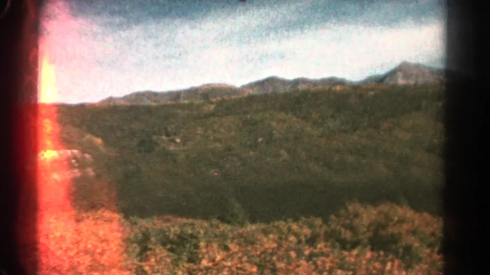 1967 Colorado mountain view with green hills, distant peaks, cloudy sky, foreground vegetation, and red film-edge light leak.