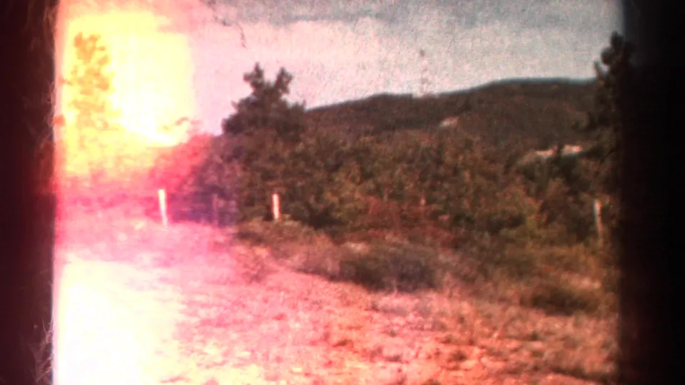 1967 Colorado 8mm frame showing a bright flared dirt path, roadside brush, fence posts, and wooded hills in the background.