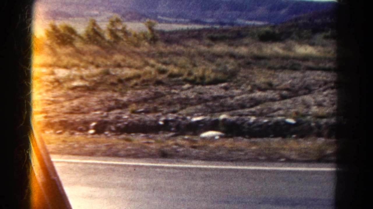 Paved roadside, dry brush, rocks, and distant hills viewed from a car window.