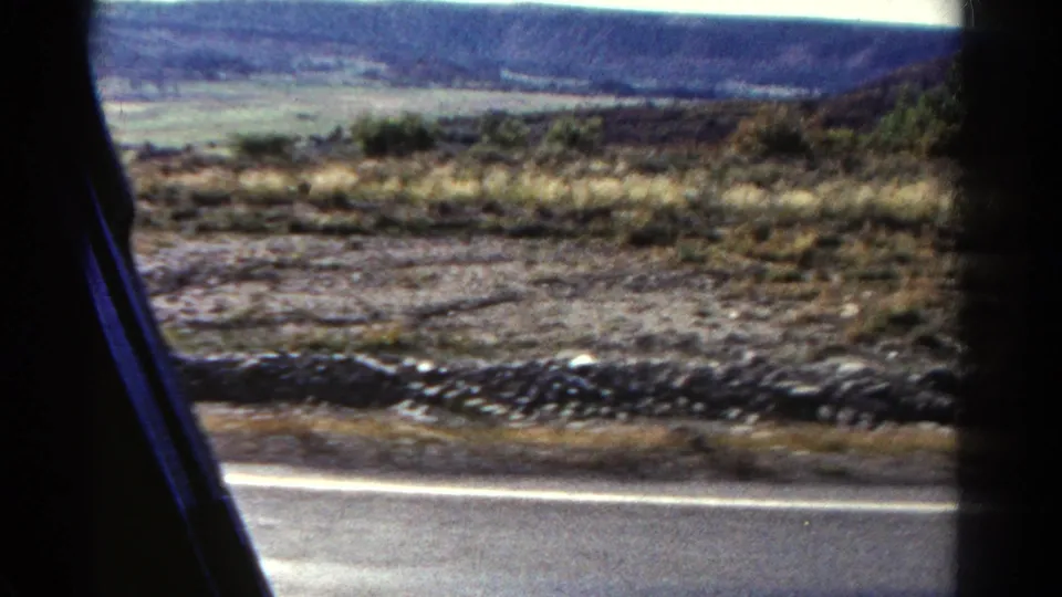 Passenger view across a Colorado roadside with scrub, stones, road edge, distant hills, and blue sky on 8mm film.
