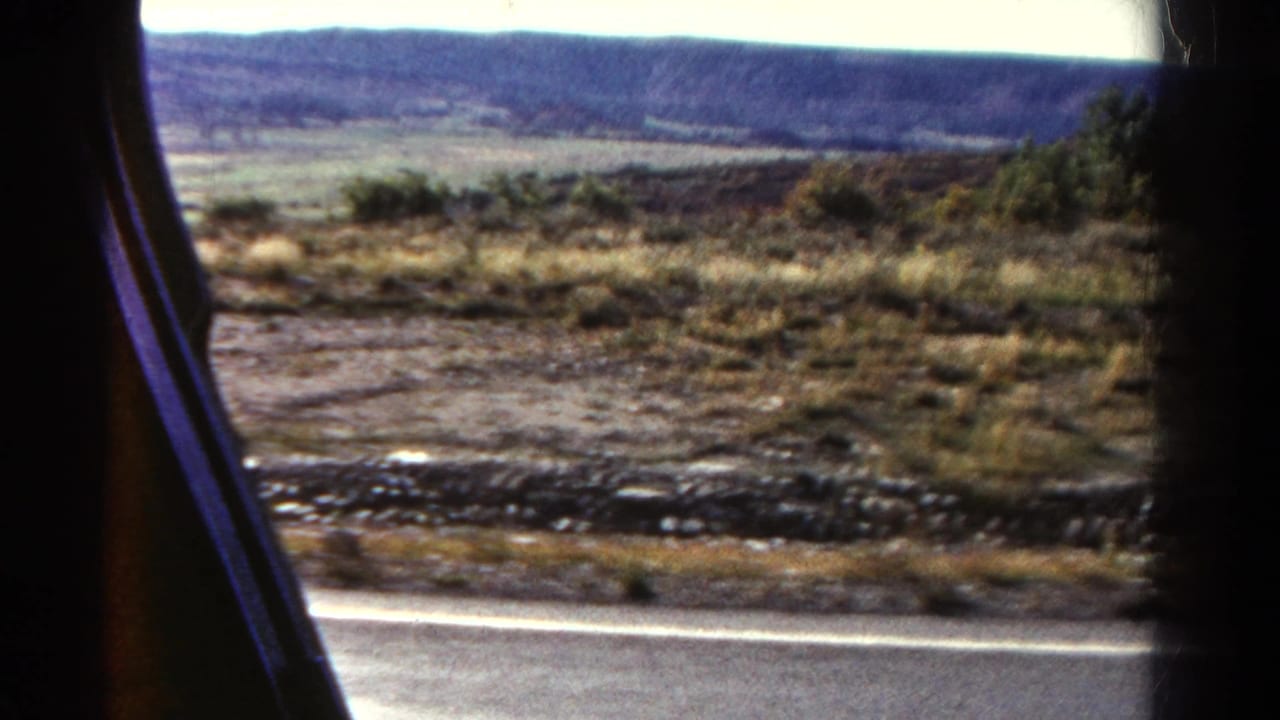 1967 Colorado road-trip view from a moving vehicle, showing a highway edge, scrubby dry ground, low stone border, and distant hills.