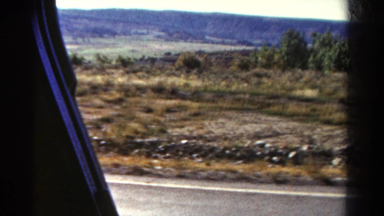 Moving 8mm view across Colorado roadside scrub, with a paved road edge in front and broad hills across the horizon.