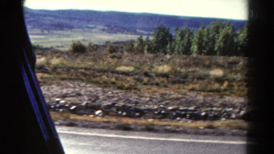 1967 Colorado highway-side landscape with a pale road stripe, rocky roadside, low vegetation, and hazy hills beyond.