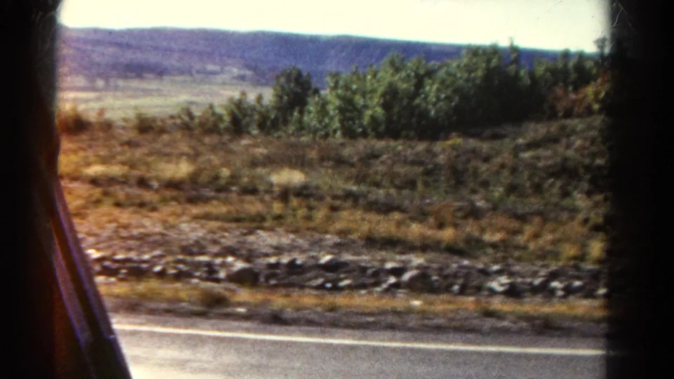 Colorado road-trip frame from 1967 showing sunlit dry vegetation, roadside stones, highway edge, and distant ridgeline.